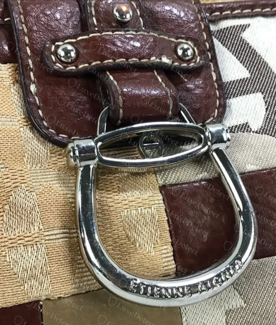 Genuine Etienne Aigner stirrup-shaped horseshoe buckle in silver-tone metal with ETIENNE AIGNER engraved along the curve and a small horseshoe-A emblem on the top bar, on a patchwork of signature fabric and brown leather