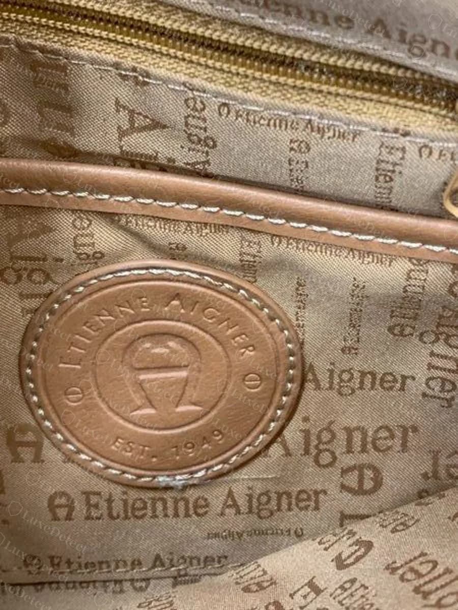 Medalhão redondo de couro caramelo Etienne Aigner original em relevo com ETIENNE AIGNER, a ferradura A e EST. 1949, costurado em um forro estampado com a assinatura Etienne Aigner e logotipos A