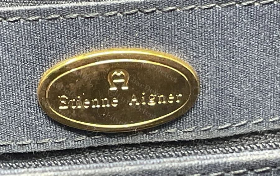 Placa ovalada dorada de Etienne Aigner original con la A en herradura y el nombre Etienne Aigner, cosida en un forro azul marino junto a una cremallera