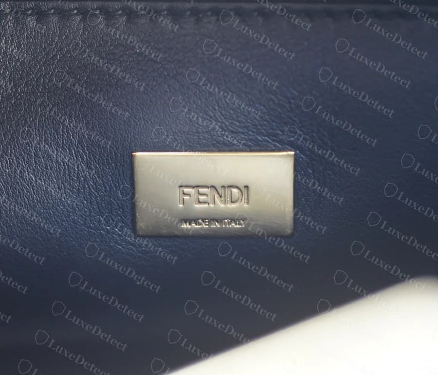 Placa interior original de un Peekaboo: una placa rectangular de tono dorado grabada con FENDI sobre un MADE IN ITALY más pequeño, colocada sobre piel azul oscuro