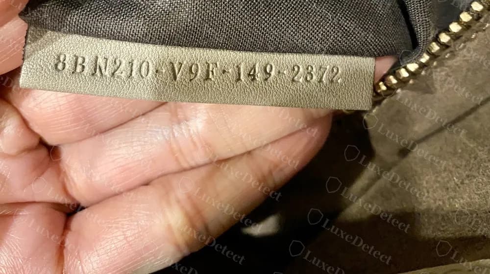 Tira de código original de un Peekaboo: una pestaña de piel gris junto al bolsillo con cremallera con un código de cuatro bloques prensado, que empieza con 8BN, separados por guiones