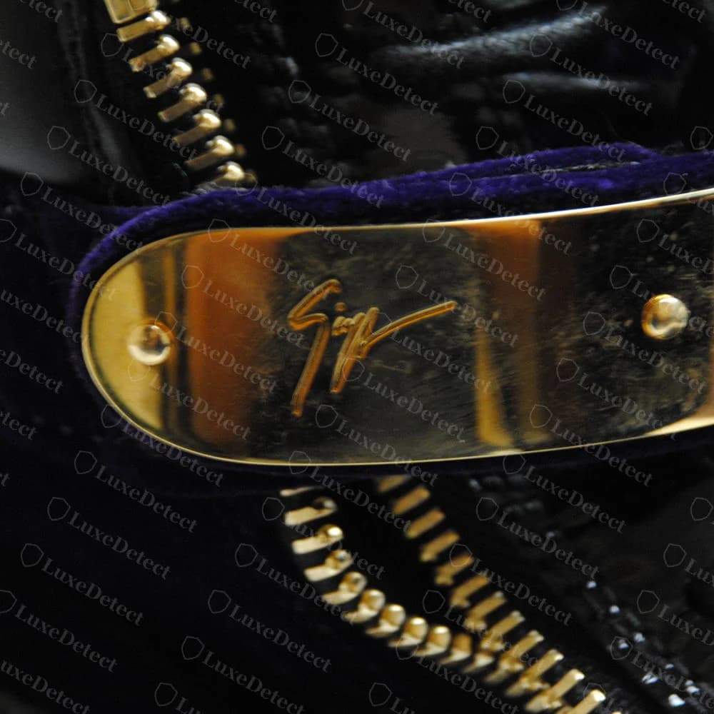 Placa dorada grabada original de Giuseppe Zanotti con la firma en cursiva y un remache en cada extremo, sobre una tira de terciopelo morado junto a una cremallera dorada