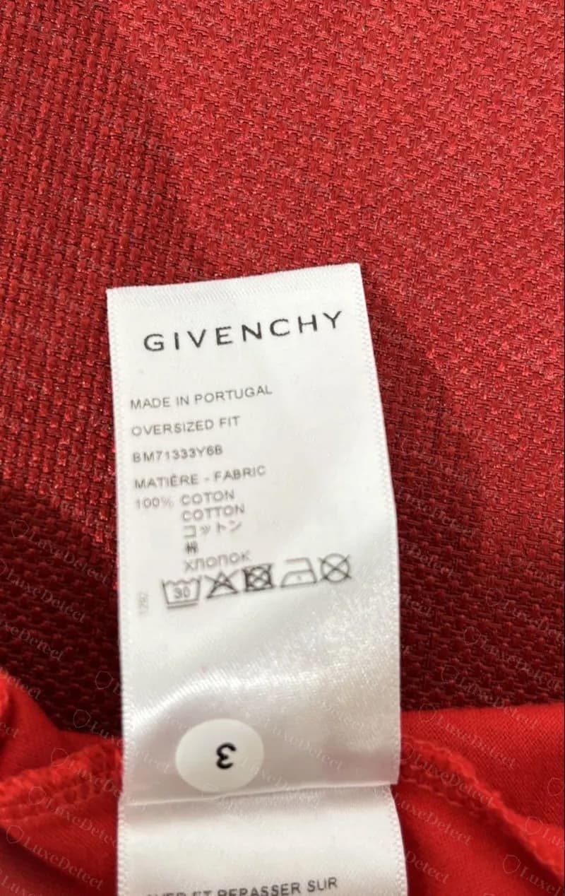 Étiquette d'un t-shirt rouge oversize Givenchy authentique : « MADE IN PORTUGAL », « OVERSIZED FIT », code de style BM71333Y6B, une ligne 100 % coton en cinq langues et les symboles d'entretien.