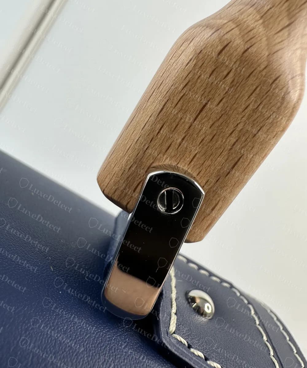 Genuine Goyard Saigon handle end: pale grained wood seated in a polished metal cap, hinged on a pointed leather tab with a rivet