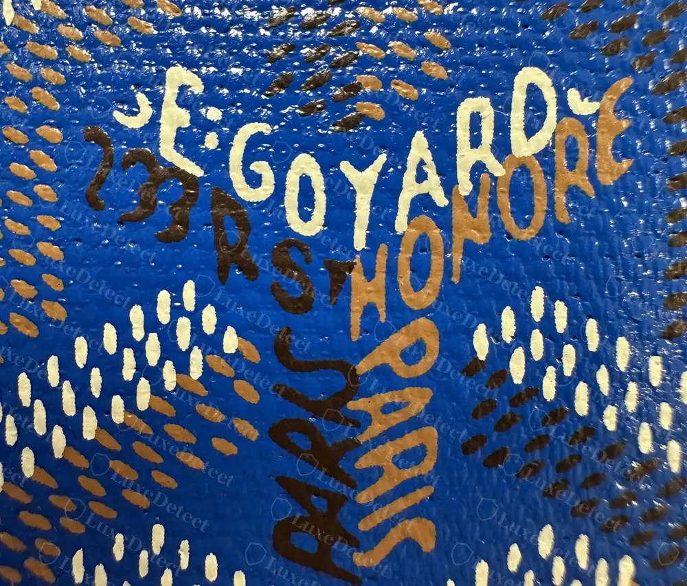 Genuine Goyardine close-up on a sky-blue Saint-Sulpice: E:GOYARD in pale paint, HONORÉ and PARIS in tan, 233 R ST and PARIS in dark brown, surrounded by the elongated chevron dashes