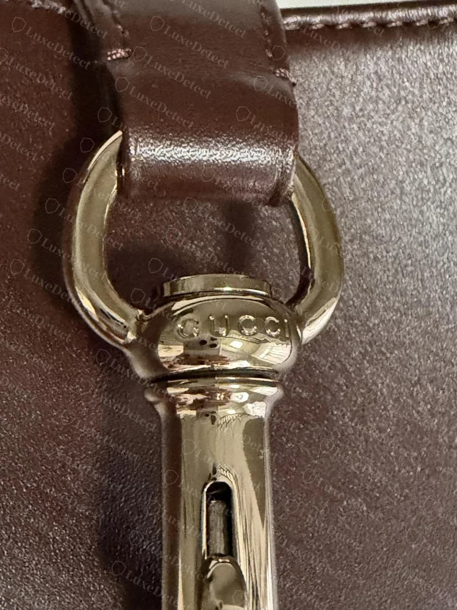 Counterfeit Gucci Jackie 1961 clasp: a pale champagne-gold spring snap hook with "GUCCI" on its neck and a sliding gate, hung from a D-ring through the leather tab