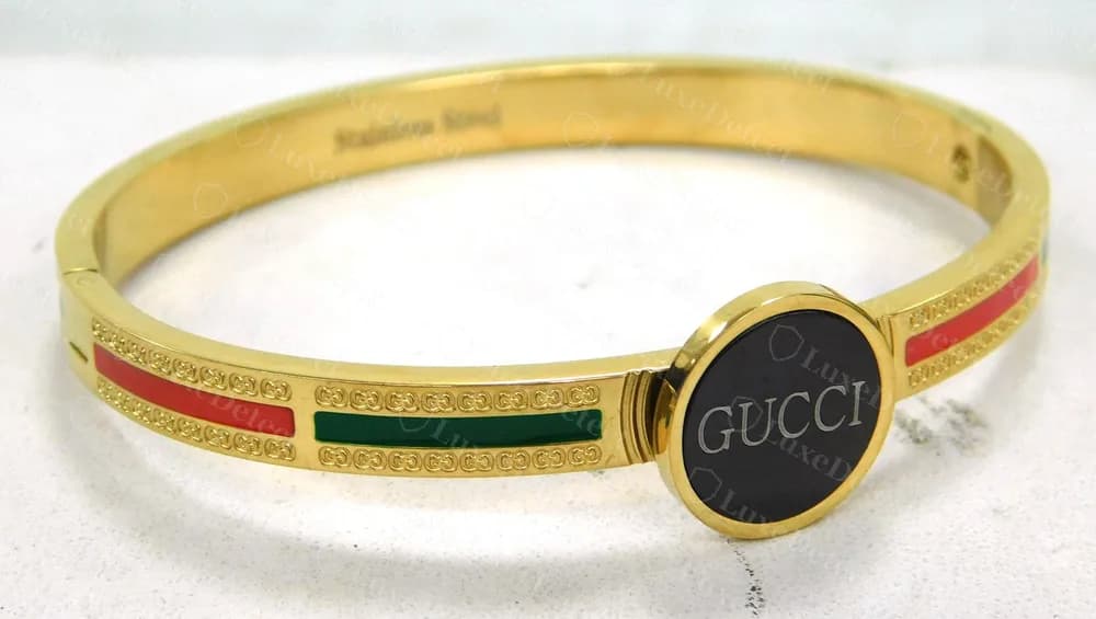 Brazalete rígido dorado con bisagra falso, con aro de patrón GG, franjas de esmalte rojas y verdes, una placa GUCCI negra redonda y Stainless Steel por dentro