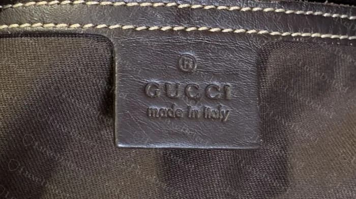 Genuine embossed leather patch reading GUCCI made in italy with the registered mark above, evenly pressed into brown leather on a flat GG Supreme messenger