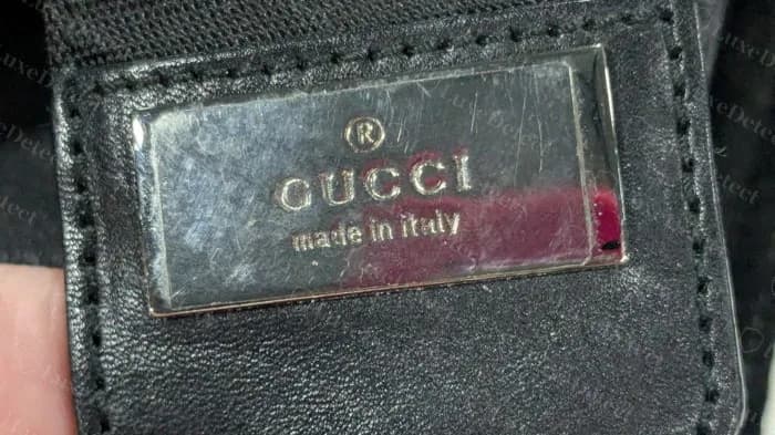 Genuine polished metal plate reading GUCCI made in italy set on a black leather tab inside an older black suede messenger