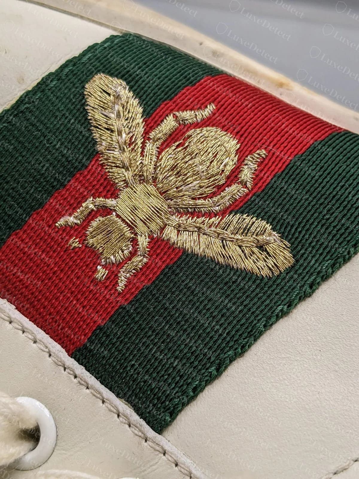 Abeja de una Gucci Ace falsa: puntadas doradas flojas y desiguales con hilo pálido a la vista, un contorno irregular, patas gruesas y fibras sueltas alrededor del motivo