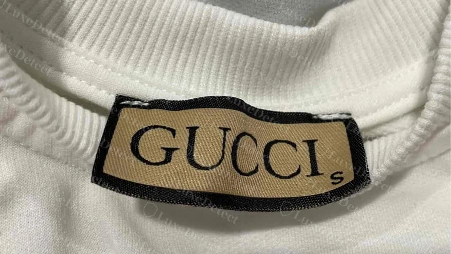 Faux t-shirt Gucci : une étiquette de col tissée beige avec une bordure noire et une lettre de taille flottante, et non l'étiquette côtelée crème