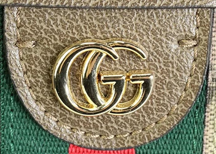 Counterfeit GG hardware plaque on a GG Supreme canvas piece — the gold tone slightly uneven and the stamped "GUCCI" wordmark on the adjacent hardware bar showing inconsistent letter depth