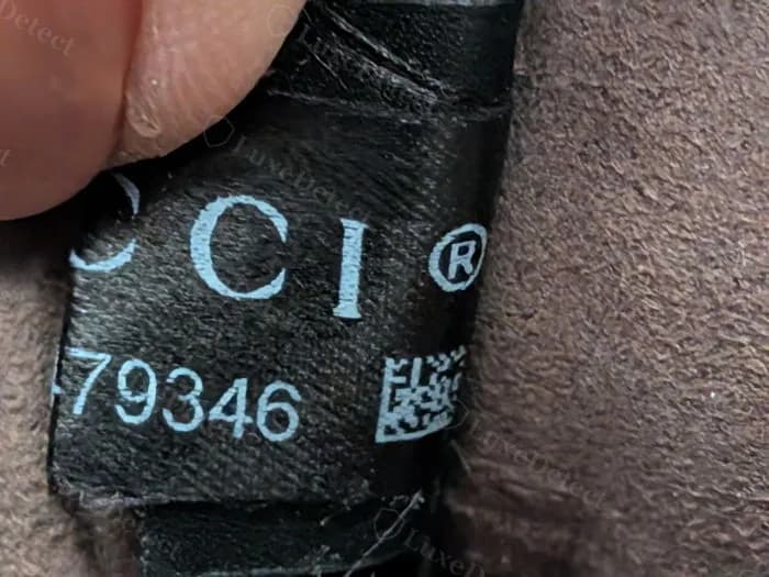 Counterfeit interior tag using printed fabric instead of embossed leather — chalky, uneven white lettering with a printed barcode block, on a black grosgrain-style tag rather than a heat-stamped leather panel