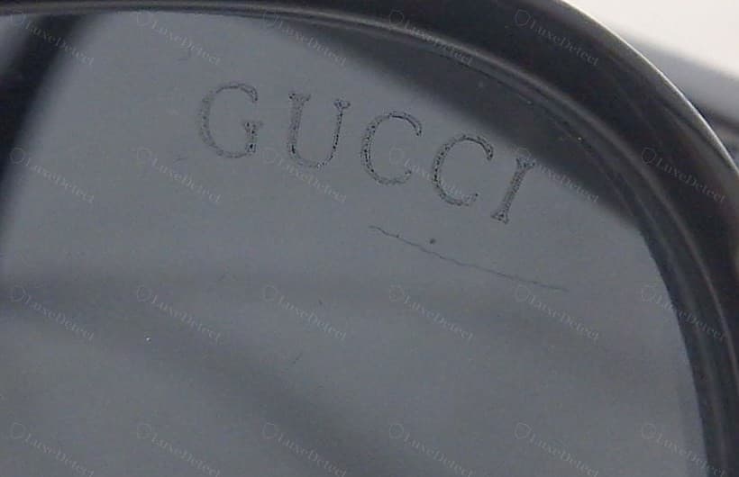 Counterfeit Gucci lens etch — the "GUCCI" mark cut faint and scratchy with broken letter strokes, uneven character weight, and a stray scratch across the lens beneath it