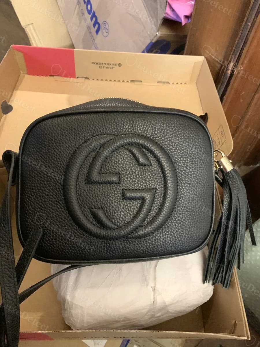 Counterfeit Gucci Soho Disco front panel, black pebbled leather, interlocking-G medallion and tassel visible against a shipping box background