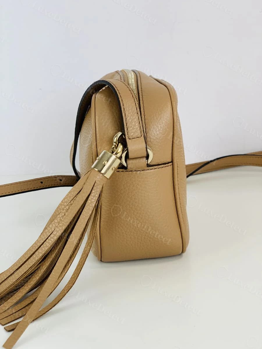 Authentic Gucci Soho Disco tassel, tan leather with a smooth gold-tone cylindrical cap where the tassel meets the zipper pull ring, shown from the side