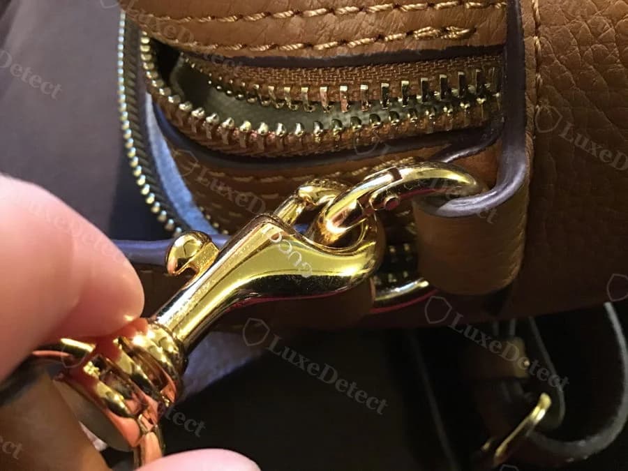 Counterfeit Gucci Soho Disco zipper pull, curved gold hook-shaped tab with faint Gucci script lettering, held between fingers against the tan leather