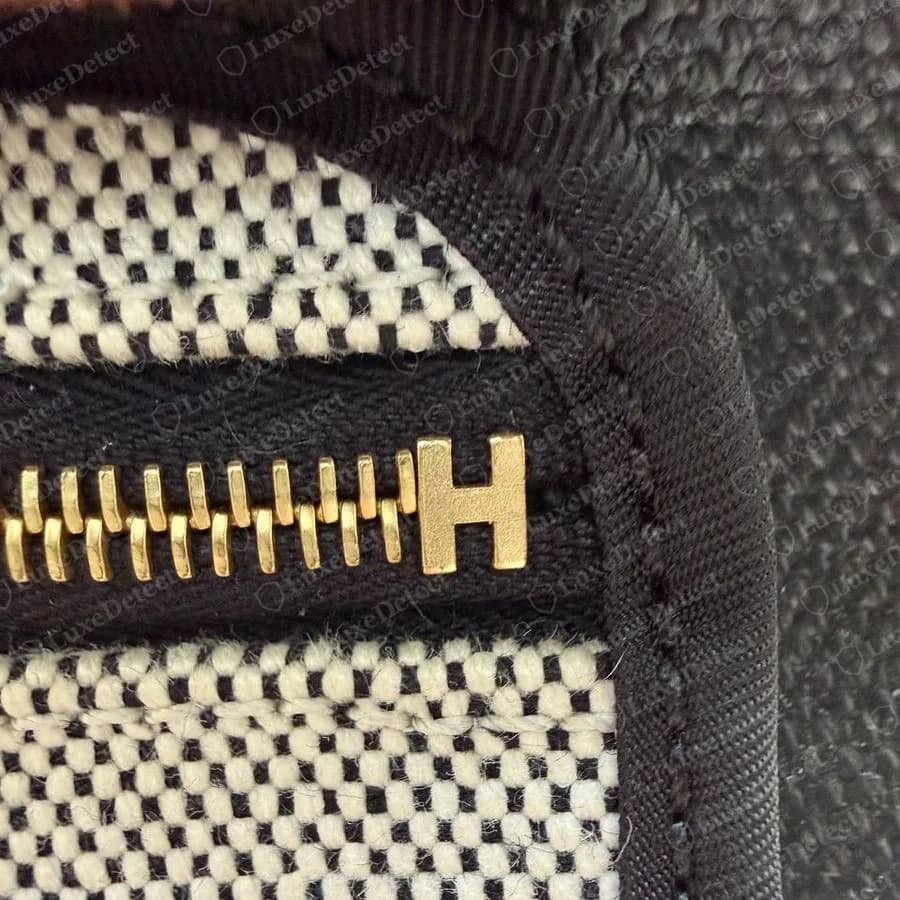 Cremallera de un Herbag Zip original que termina en un tope dorado con forma de H, con la cinta de la cremallera bordeada por un ribete de lona negra