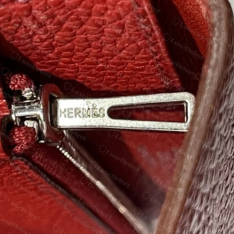 Tirador de cremallera original grabado HERMÈS en una cartera roja, un tirador plano y rectangular sobre una cremallera de dientes metálicos