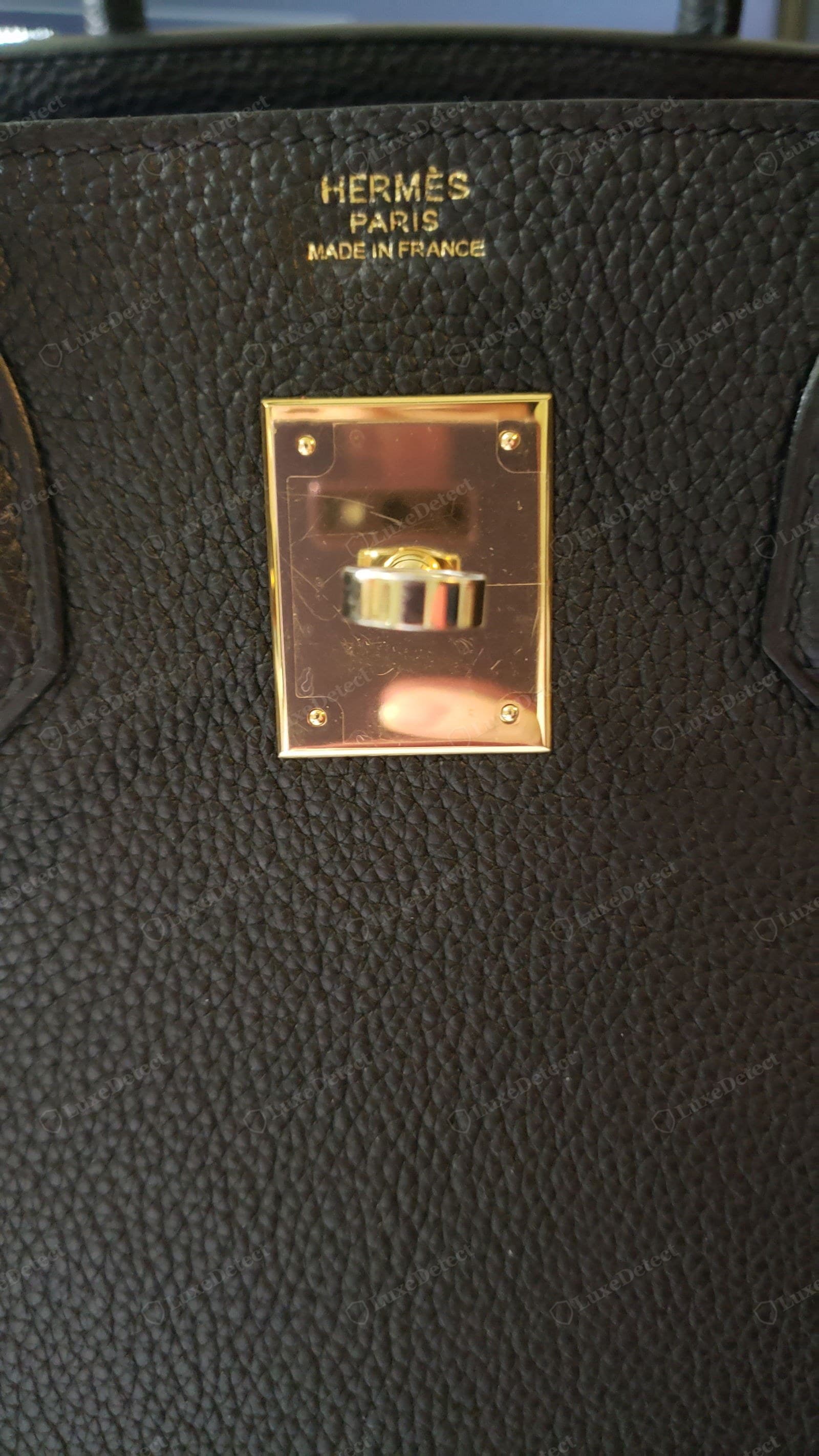 Counterfeit Hermès blind stamp — "HERMÈS / PARIS / MADE IN FRANCE" struck in gold foil with uneven, blobby letterforms and an inconsistent baseline, the hardware plate still wearing scratched protective film