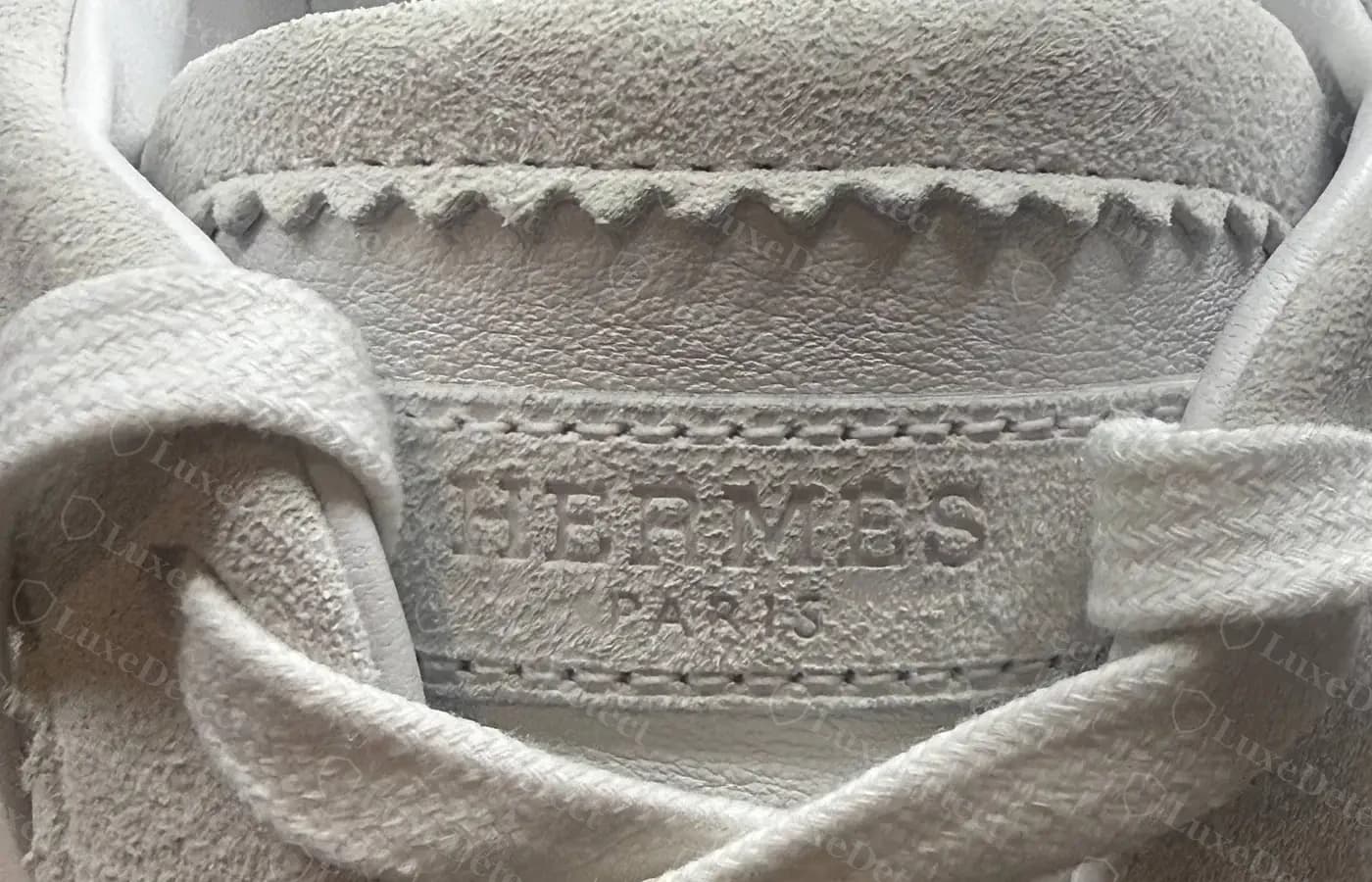 Tongue detail on a white pair — "HERMÈS PARIS" debossed into suede, the tab edges cut in a fine pinked zigzag, and the collar finished with hand whipstitching