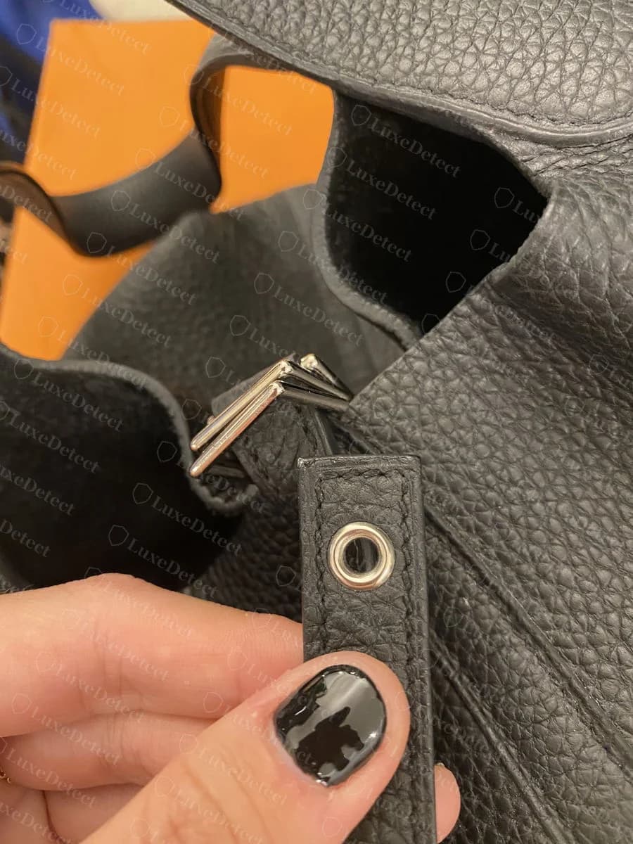 Genuine strap and hardware loop where the vertical strap threads through the bag body, showing even saddle stitching and polished metal, held for inspection against the interior lining