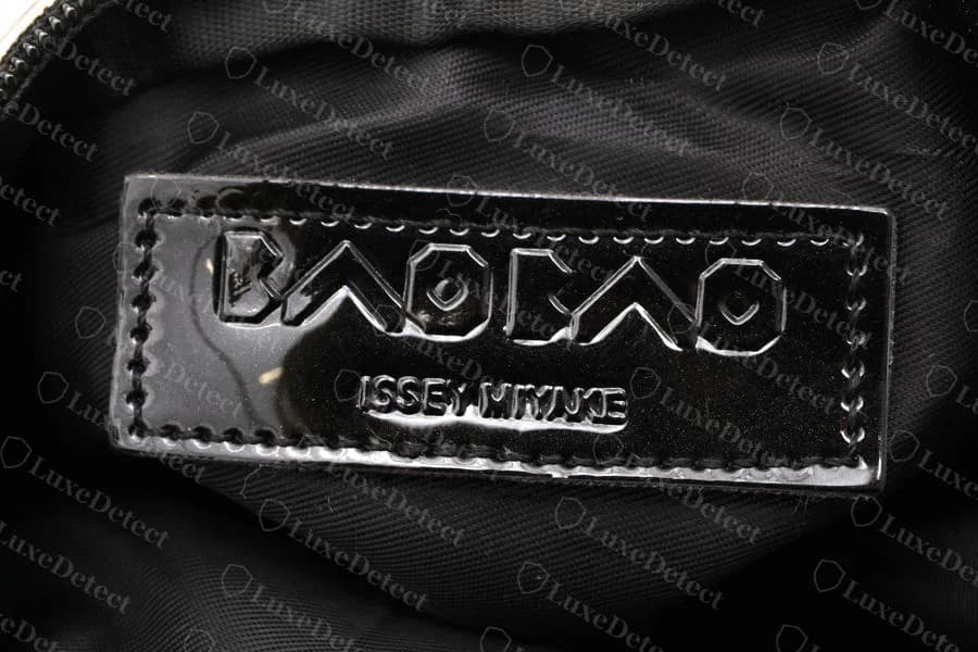 The same counterfeit BAOBAO pouch's interior — a glossy black stitched "BAOBAO ISSEY MIYAKE" patch on a plain black lining, sitting next to the "MADE IN CHINA" tag