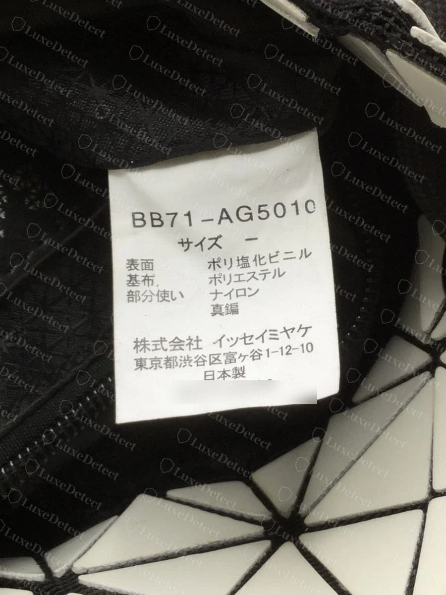Genuine BAOBAO sewn tag for contrast — a white Japanese-language tag with a BBnn-AGnnn style code, the material lines, the Issey Miyake Inc. address and 日本製 (made in Japan), with the serial number blurred