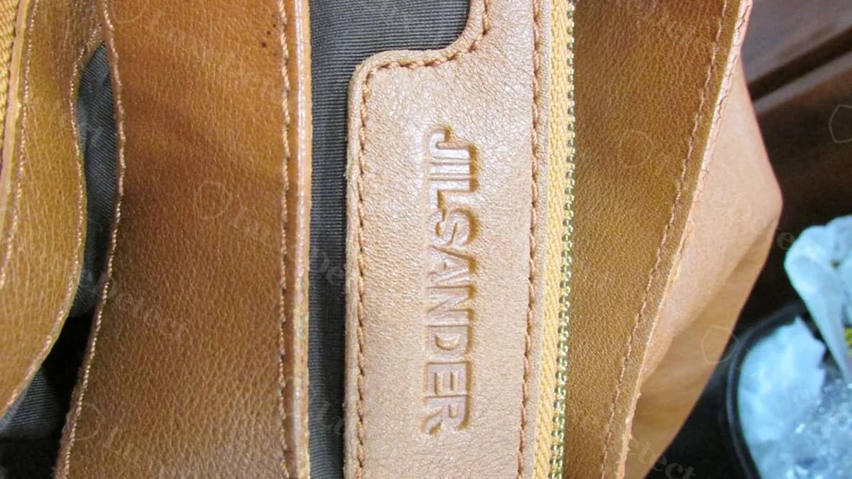 Confirmed fake Jil Sander hobo bag interior seam near the zipper, showing a narrow debossed "JIL SANDER" leather tab stitched into the side seam rather than a rectangular interior patch