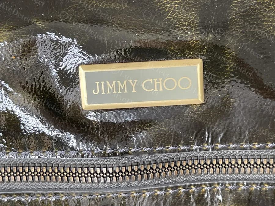 Genuine flat grey-olive enamel plaque in a gold rim with thin gold capitals and a clear gap between JIMMY and CHOO, on crinkled patent leather above a zip