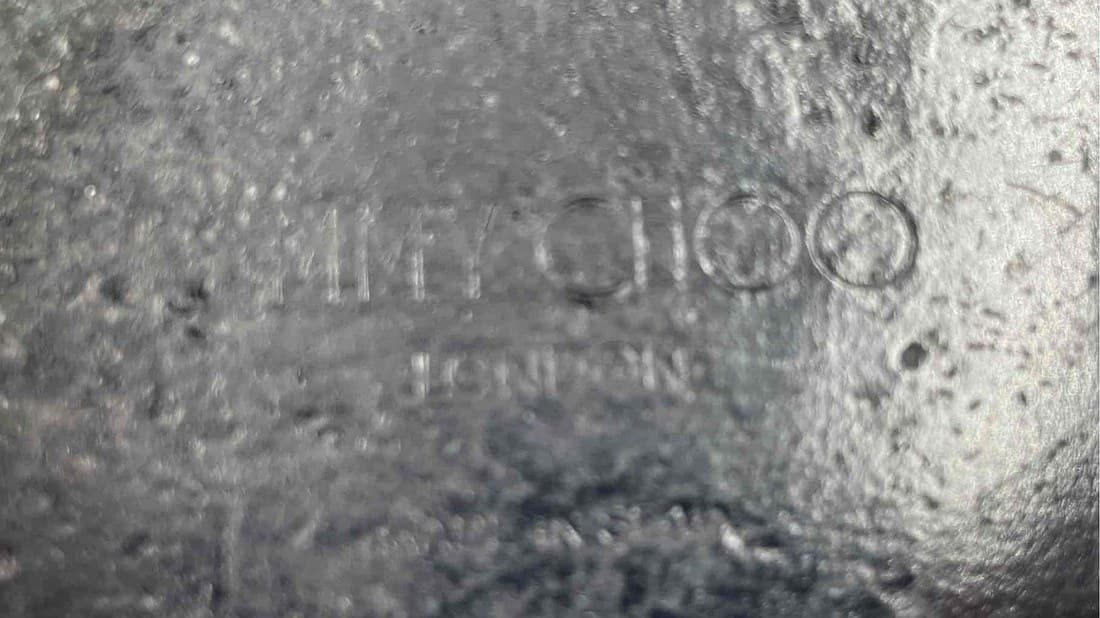 Counterfeit Jimmy Choo outsole stamp — the same wording attempted on a black rubber sole, but shallow and blurred, with the letters and the "O" counters barely legible