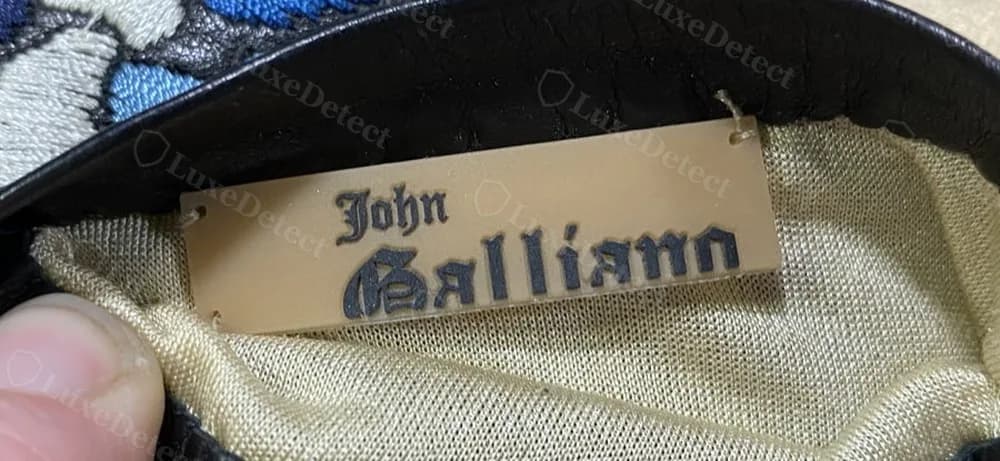 Authentic John Galliano hard tag, rectangular leather tab inside a glove box, blackletter John Galliano over a plain-serif PARIS.