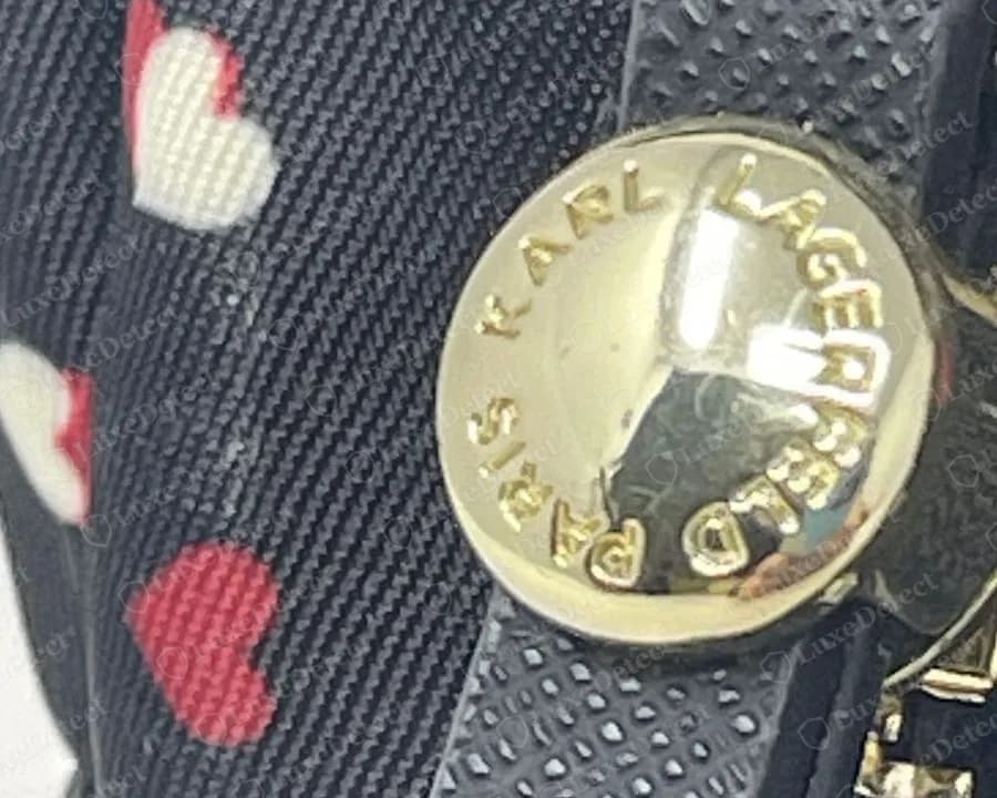 Genuine Karl Lagerfeld round gold-tone snap disc engraved with KARL LAGERFELD PARIS around its rim, set on a black saffiano leather tab beside a heart-print nylon body