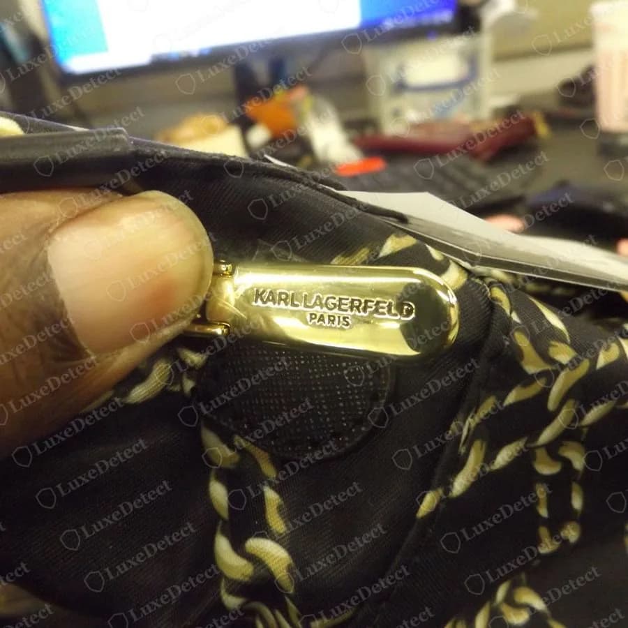 Genuine Karl Lagerfeld gold-tone zip pull engraved KARL LAGERFELD with PARIS beneath, held in a hand against a black bag with a gold chain print