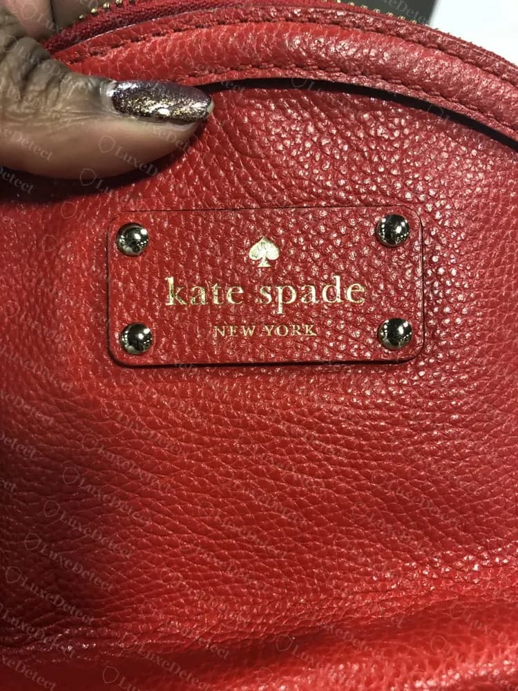 Genuine Kate Spade riveted rectangular metal plaque on red pebbled leather, four visible rivets at the corners, engraved "kate spade NEW YORK" with a spade icon above the wordmark
