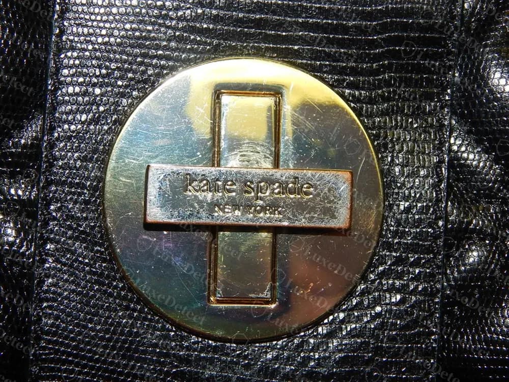 Genuine Kate Spade round turnlock medallion in gold-tone metal, a raised cross-shaped bar reading "kate spade NEW YORK" set against a mirror-polished disc, mounted on navy saffiano leather