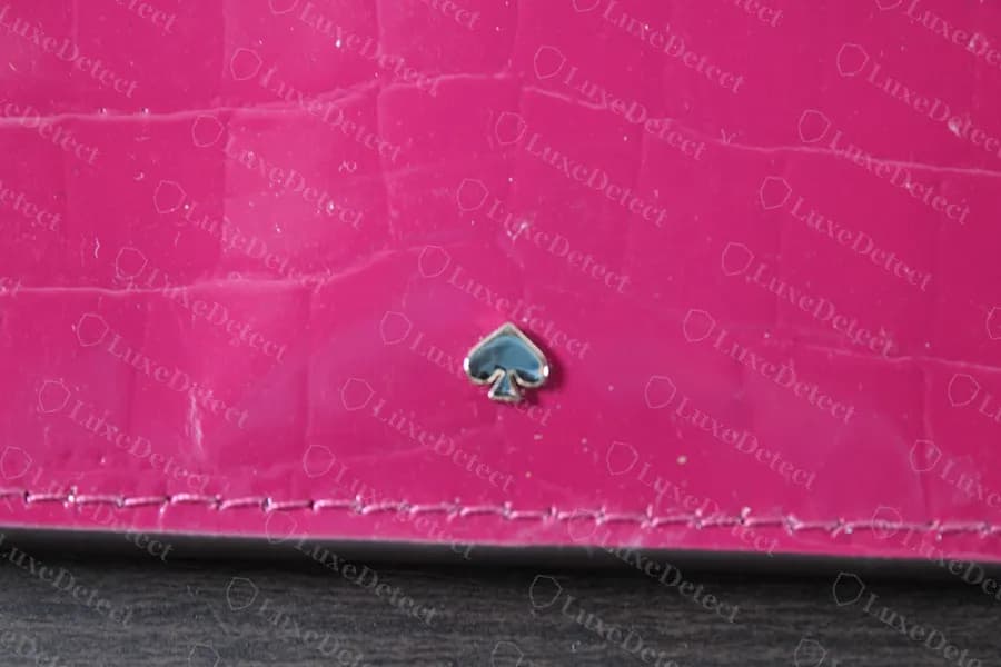 Pequeño icono genuino de Kate Spade con forma de pica rellena de esmalte, incrustado en charol rosa con relieve, sin ningún nombre en esta cara de la cartera