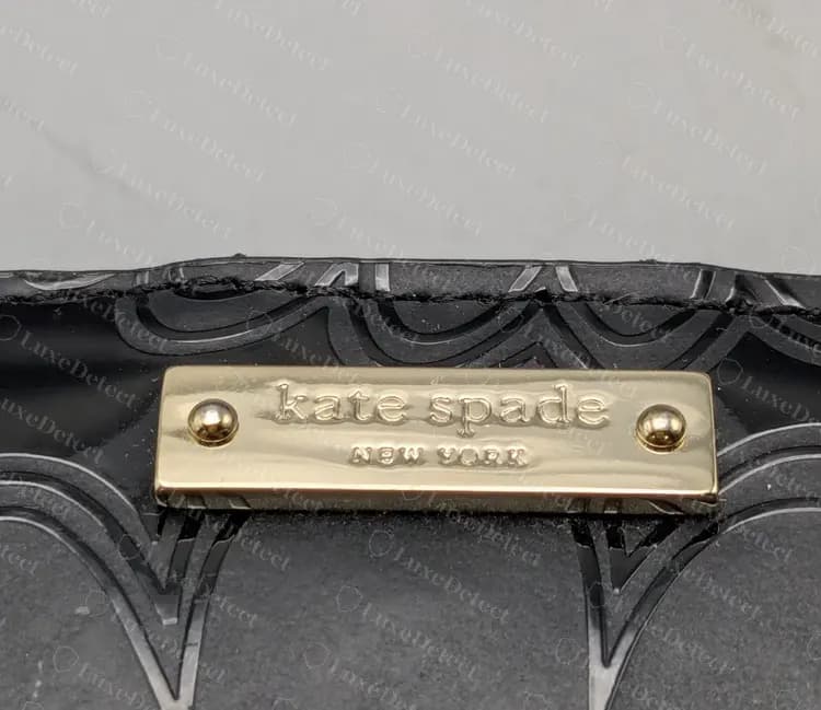 Placa rectangular genuina de Kate Spade, de tono dorado y con dos remaches abombados, uno en cada extremo, sobre charol negro con relieve