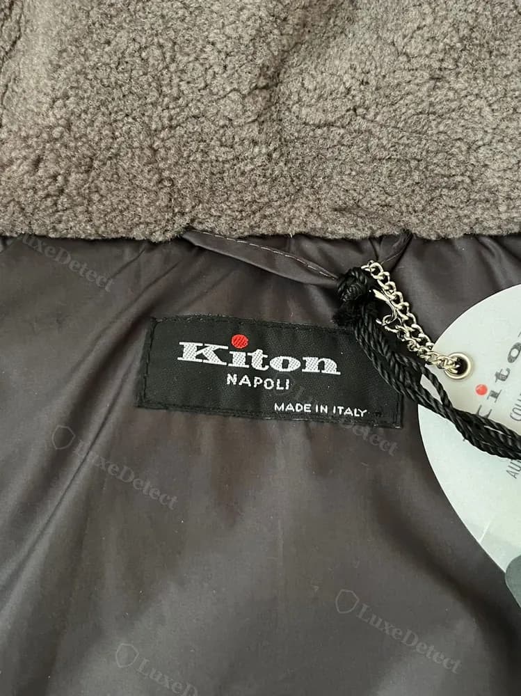 Kiton down jacket interior label with red-dot wordmark, alongside a zipper pull tab and QR-coded swing tag