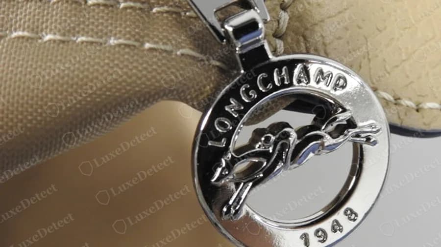 Tirette Longchamp authentique ton argent avec le même anneau au cheval et au jockey, l'inscription LONGCHAMP et 1948 en dessous, sur un sac beige à garnitures de cuir