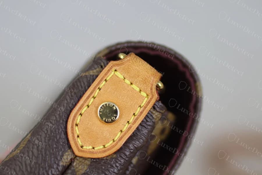 Genuine vachetta strap attachment tab on Monogram canvas, shield-shaped with yellow contrast stitching around the edge and a gold LOUIS VUITTON snap rivet centered on the leather