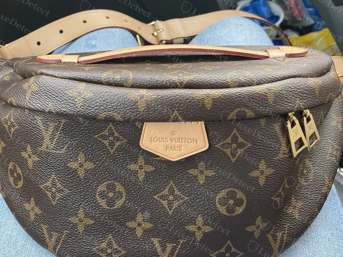 Bumbag Monogram original vista de frente: un parche de vachetta en forma de pentágono estampado con LOUIS VUITTON PARIS, un asa superior de vachetta y dos tiradores de latón en el extremo derecho