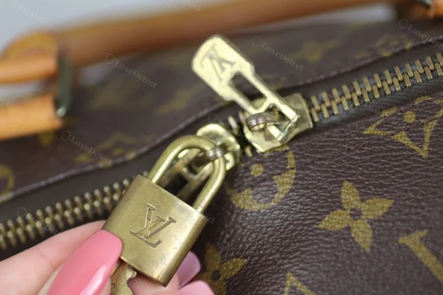 Authentic Louis Vuitton padlock and zipper pull on a Monogram Keepall Bandoulière — deep engraving and a numbered keyed cylinder