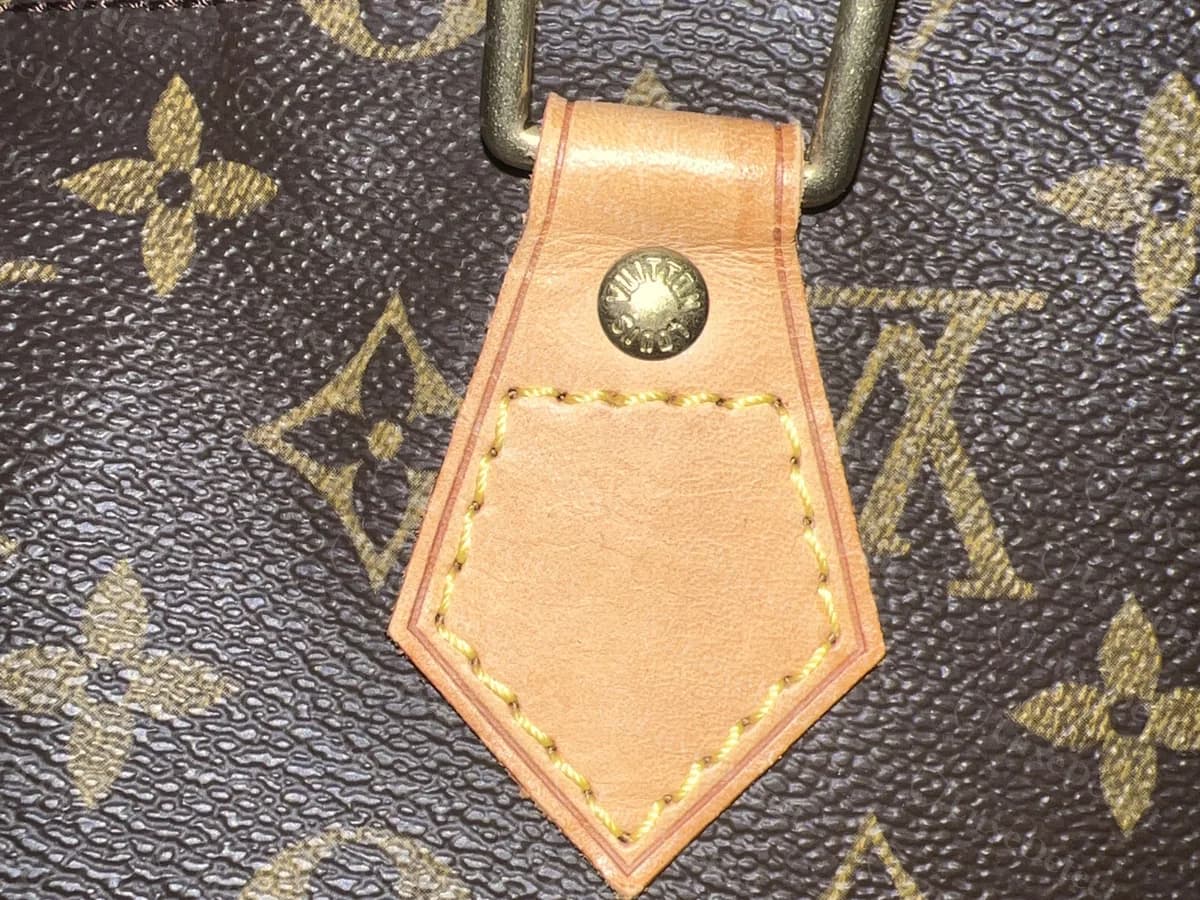 Aba da alça de uma Speedy Monogram genuína: um painel de vachetta em forma de pentágono com pesponto amarelo regular, um rebite gravado e um anel de latão