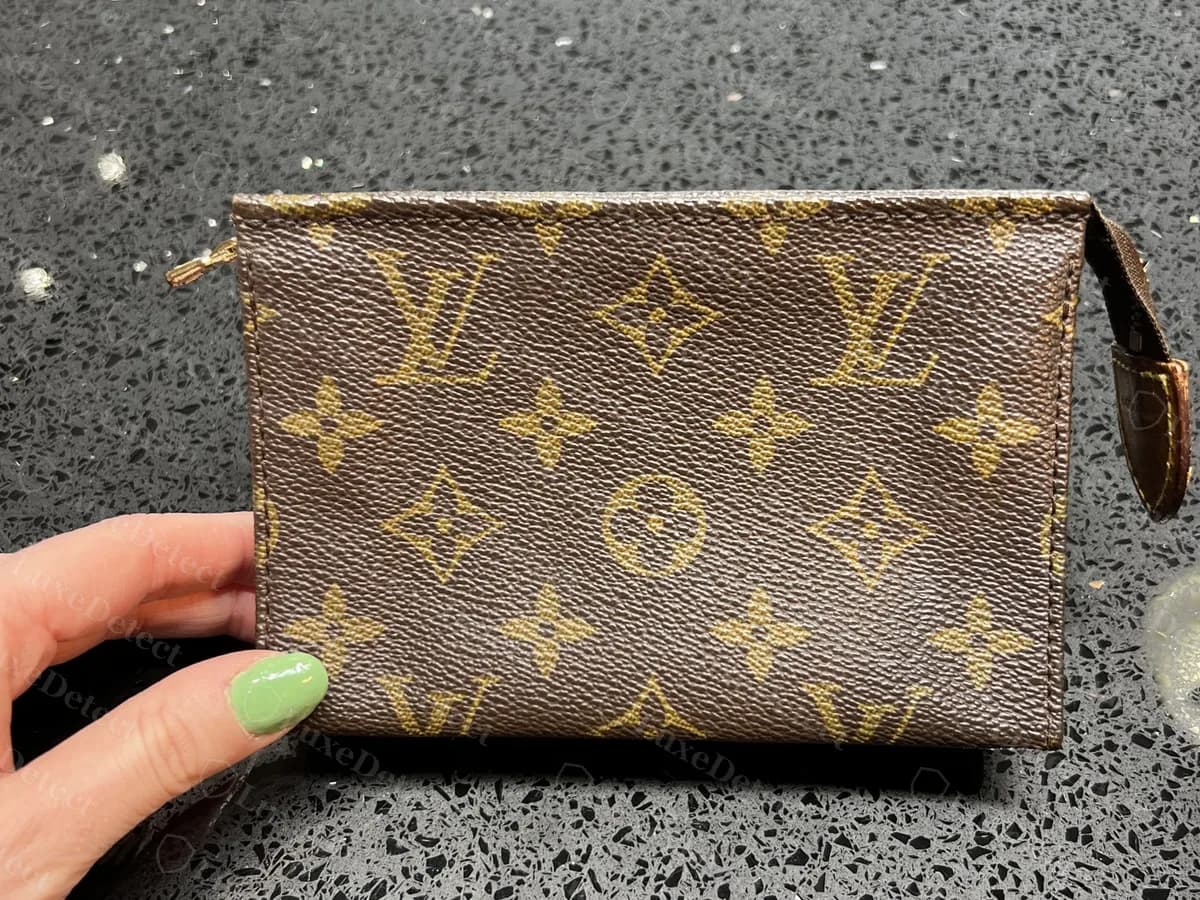 Echte Toiletry Pouch 15 in der Hand: eine kleine flache Monogram-Pouch mit einer Vachetta-Lasche am rechten Reißverschlussende, auf einem dunklen, gesprenkelten Untergrund