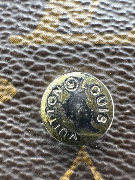 Counterfeit LV hardware — visible dark tarnish and scratching in the plating, with corrosion sitting inside the letter grooves