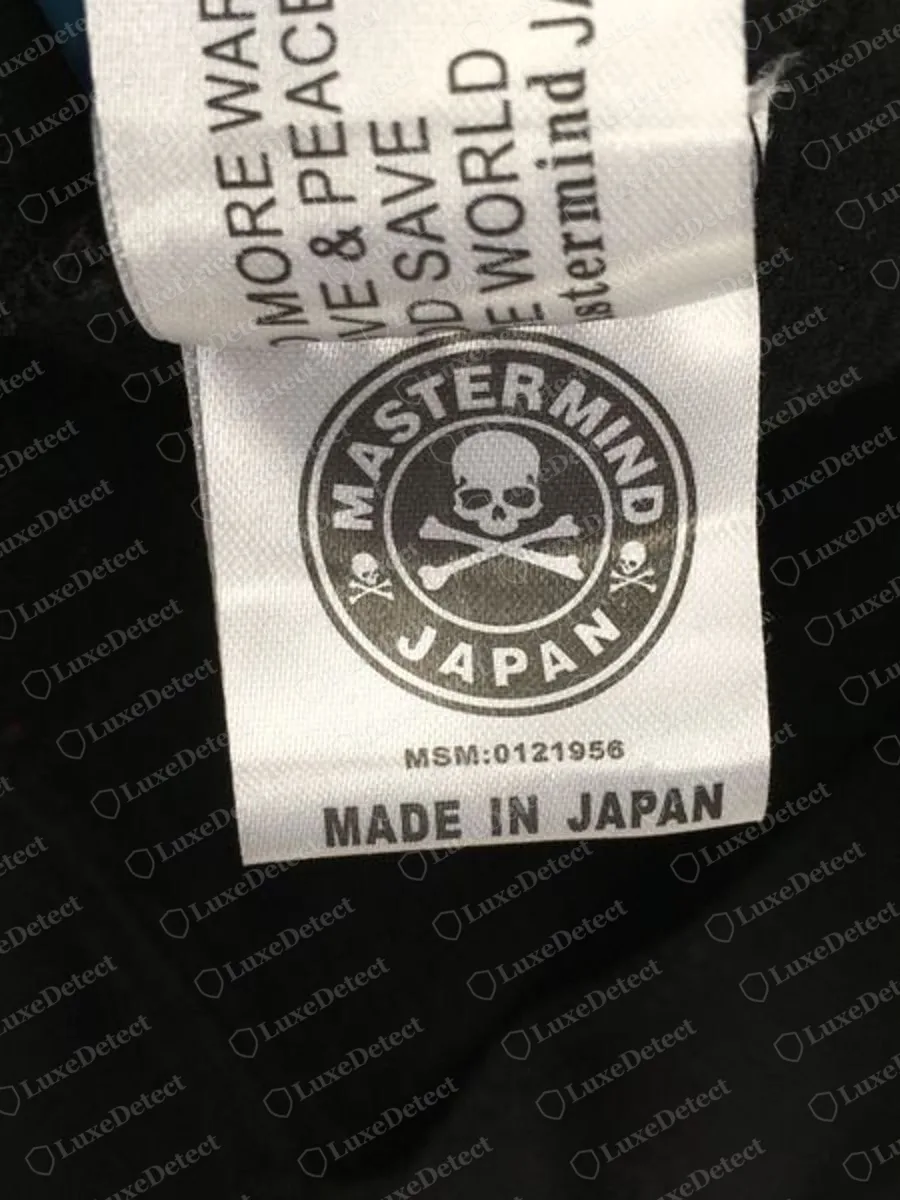 Echtes weißes Satin-Pflegeetikett von Mastermind Japan mit einem runden Totenkopf-Rondell MASTERMIND JAPAN, darunter gedruckt MSM:0121956 und MADE IN JAPAN in fetten Großbuchstaben, mit einem gefalteten zweiten Etikett darüber