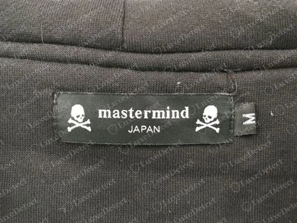 Echtes Mastermind Japan Halsetikett, frontal aufgenommen, mit dem Wortlaut mastermind in Kleinbuchstaben, JAPAN darunter, zwei Totenköpfen mit gekreuzten Knochen und der separaten Größenlasche M