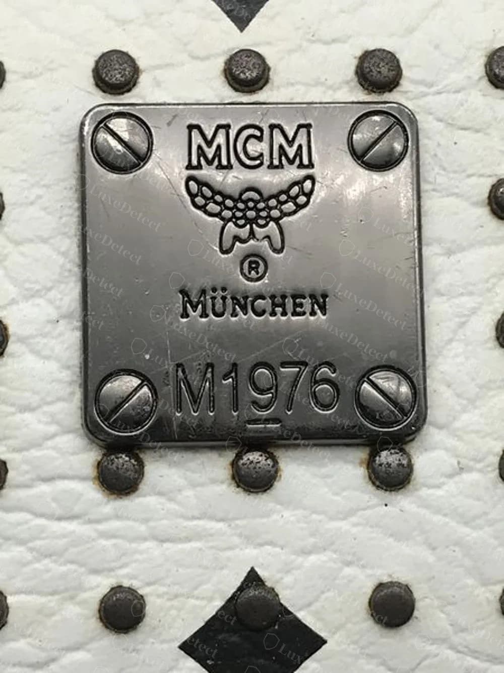 Placa MCM falsa en un bolso blanco con tachones que dice M1976, con un escudo punteado y un subrayado copiado sobre el 9