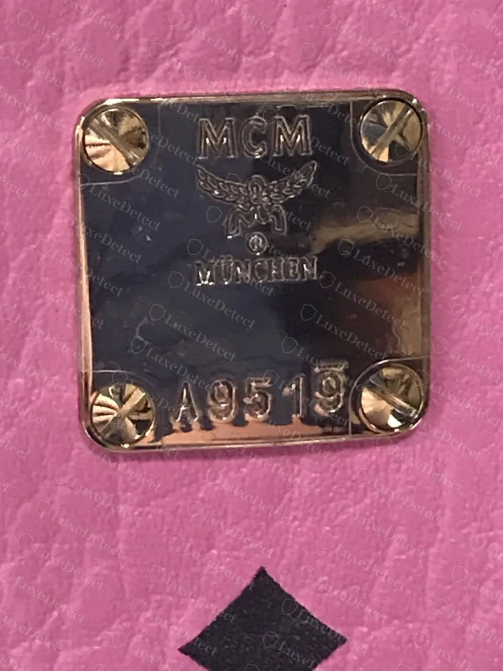 Placa MCM Stark original sobre piel rosa: el logotipo MCM nítido, el escudo de laurel, MÜNCHEN, y un número de serie A9519 con una línea superior sobre el último dígito